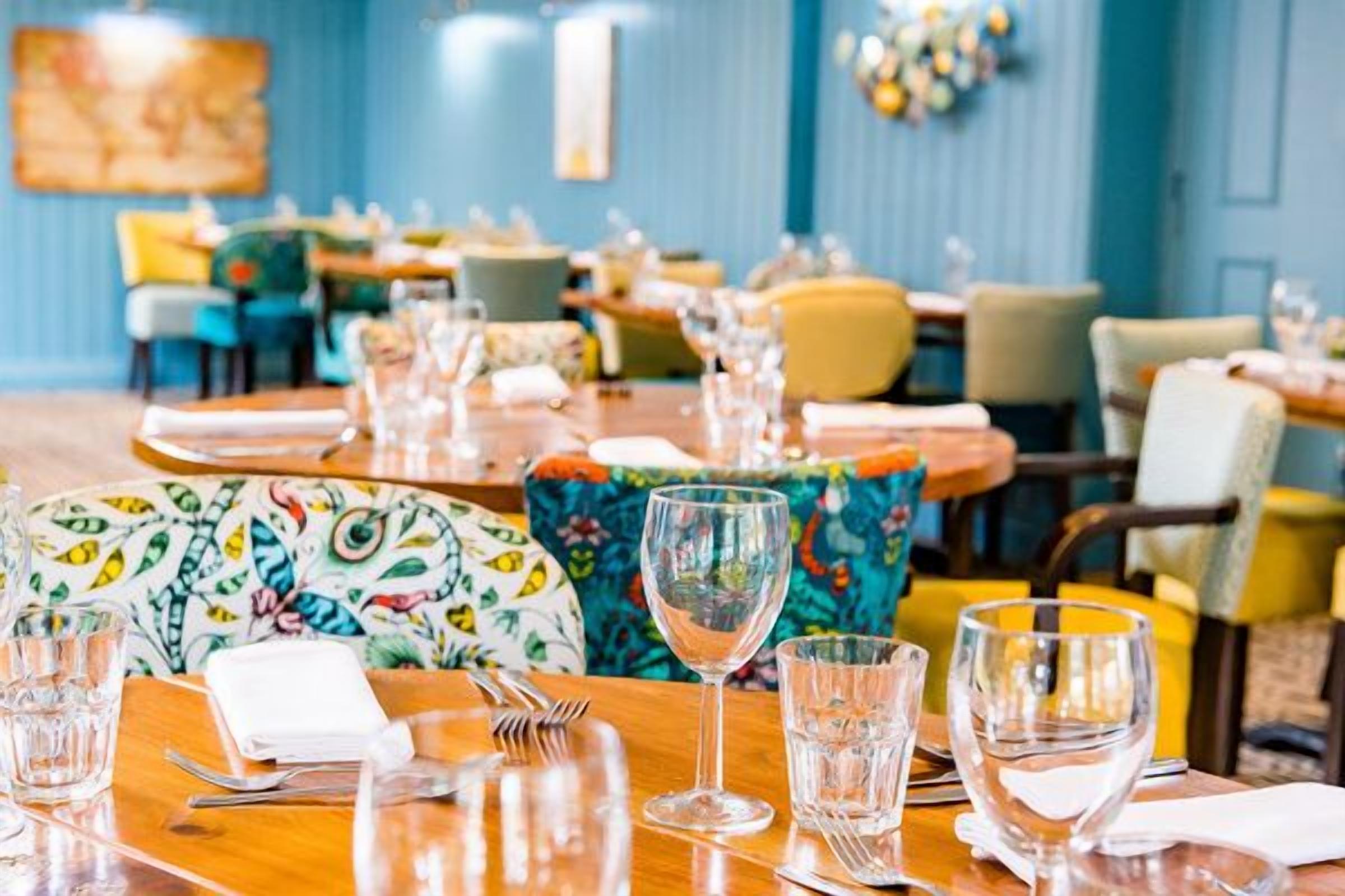 A dining area with tables set for service, chairs with colorful upholstery, and a blue wall with decorative elements.