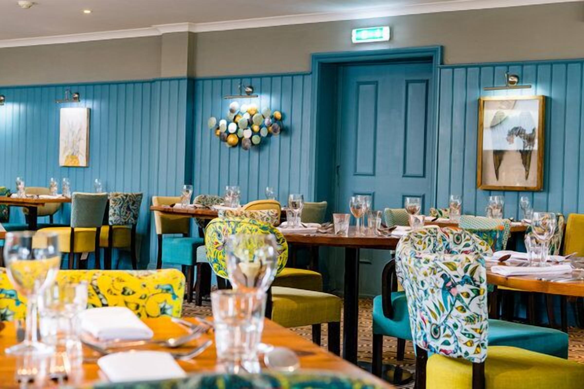 Un comedor con mesas dispuestas para comidas, sillas con tapicería colorida y una pared azul con detalles decorativos.