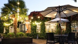 An outdoor patio area with a table and chairs under a large umbrella, surrounded by lush greenery and string lights. - Rossett Hall Hotel (Wrexham)
