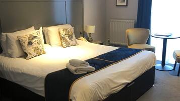 Quarto de hotel com uma cama grande, uma mesinha de cabeceira com abajur, uma cadeira e uma pequena mesa com telefone.
