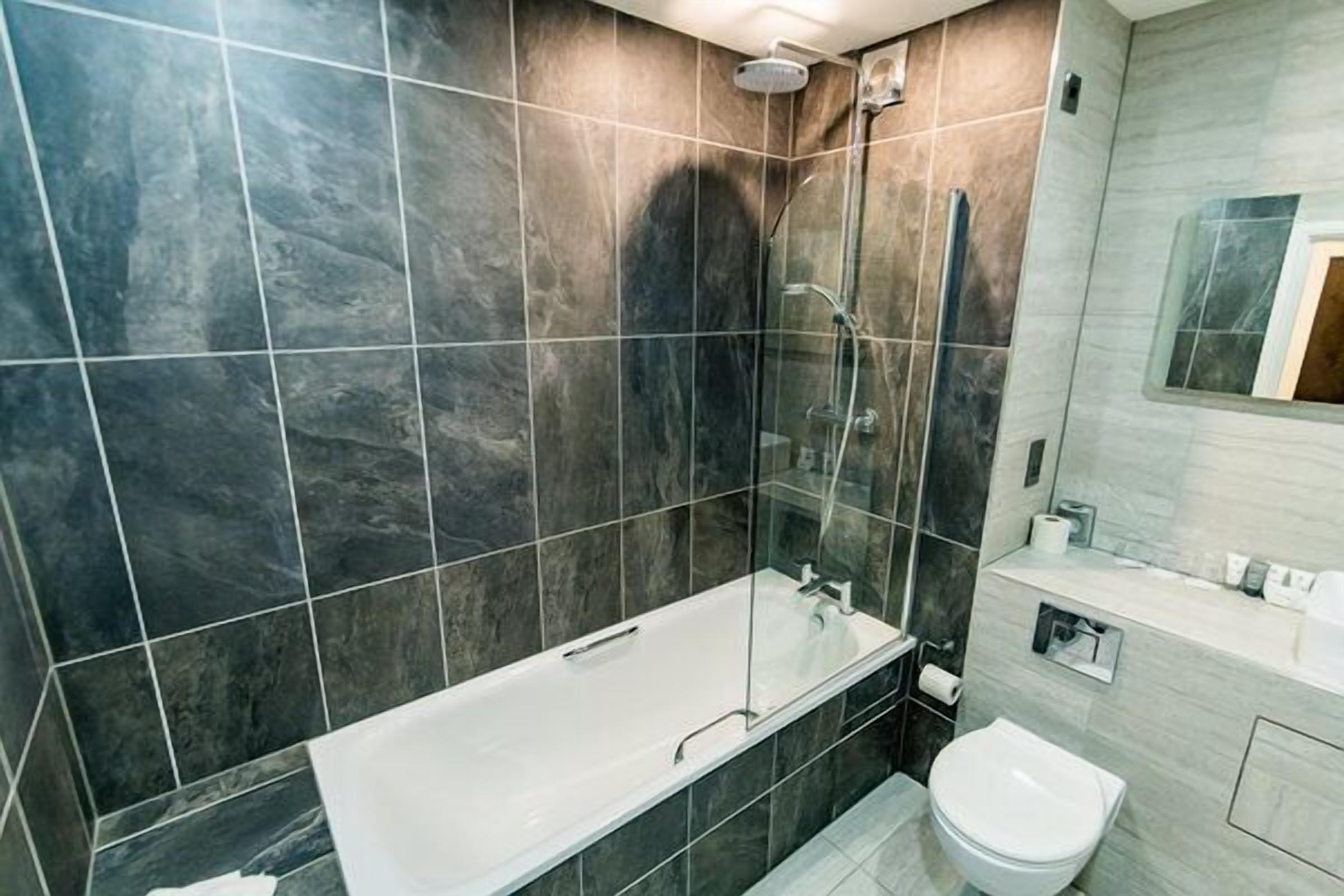 Moderni kylpyhuone, jossa on lasiseinällä varustettu suihku, valkoinen kylpyamme ja wc.