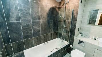 Moderni kylpyhuone, jossa on lasiseinällä varustettu suihku, valkoinen kylpyamme ja wc.