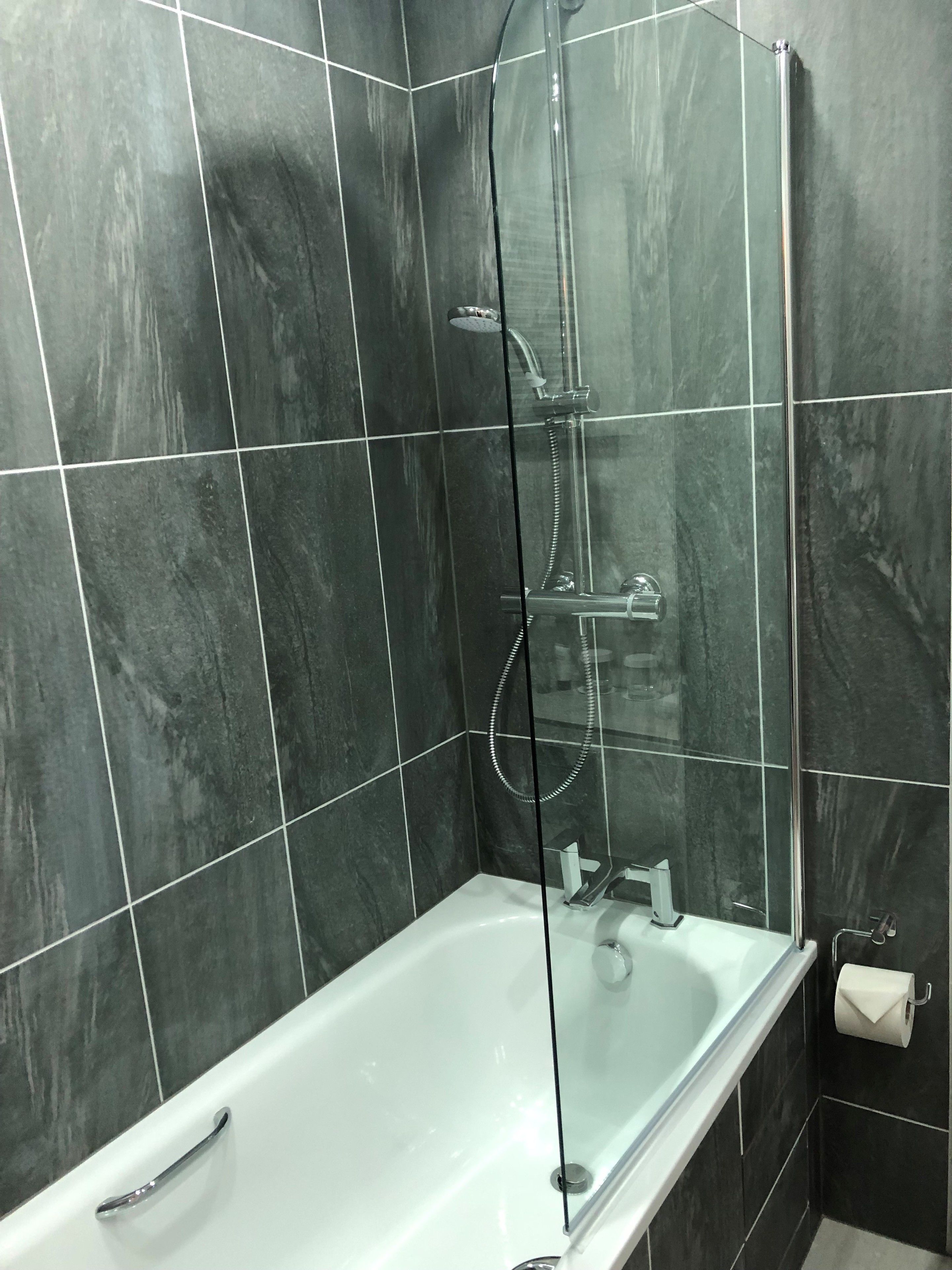 A modern bathroom with a glass-enclosed shower, a white bathtub, and dark tiled walls.