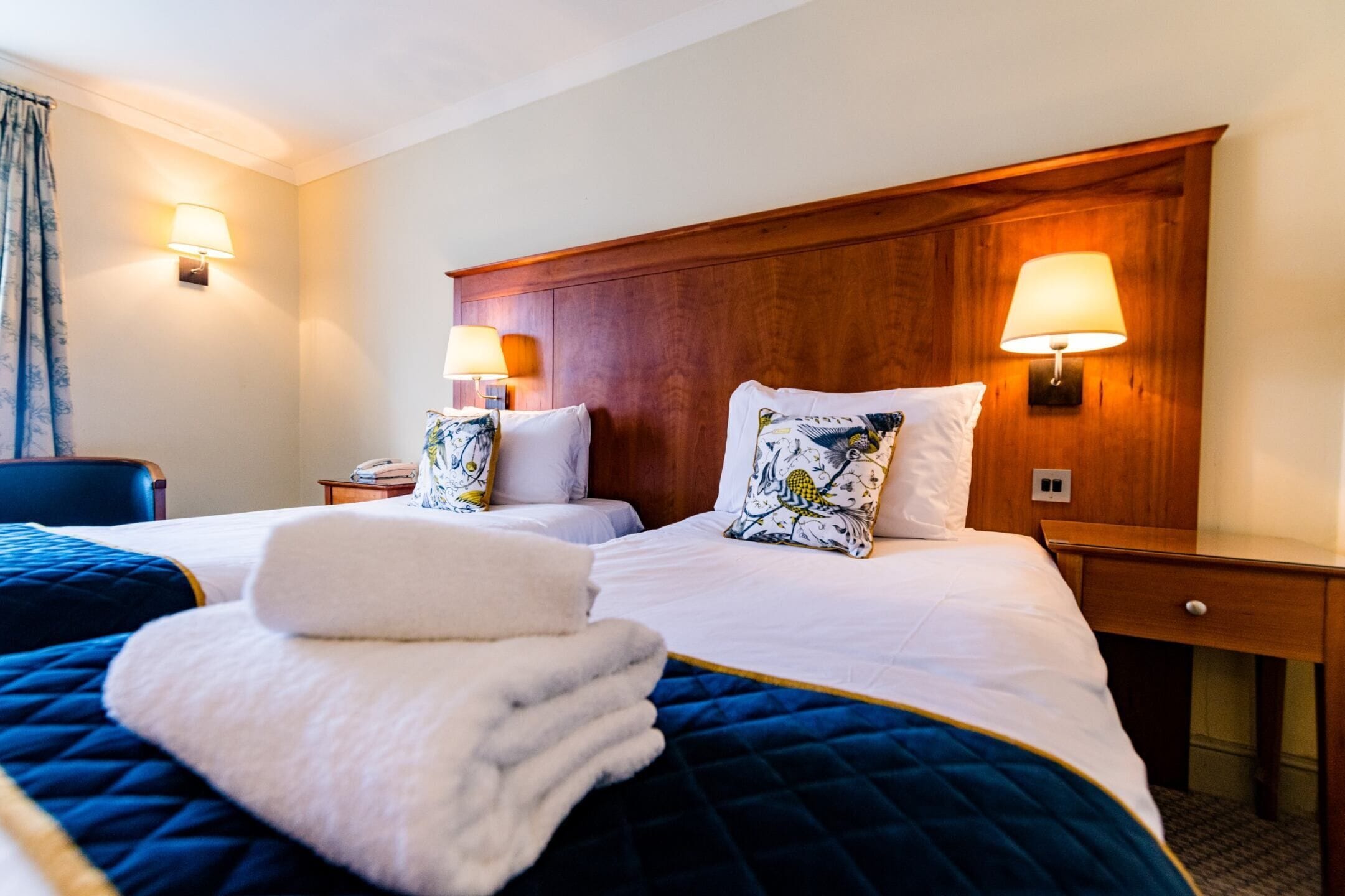 Une chambre d’hôtel avec deux lits, chacun doté d’une tête de lit en bois, de draps blancs et d’oreillers décoratifs. On y trouve des lampes de chevet et une petite table avec une lampe.
