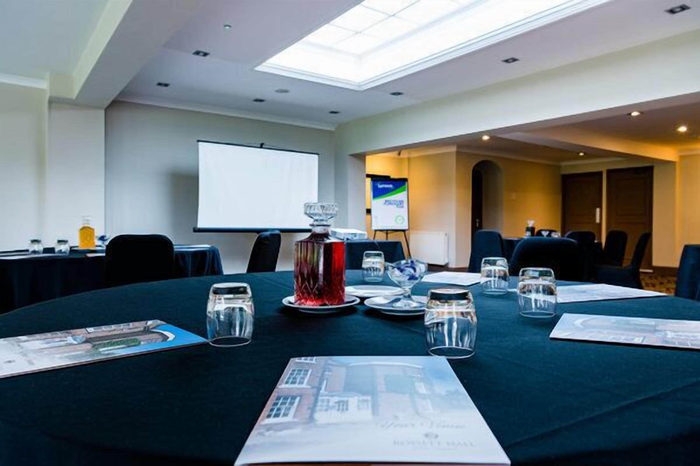 Uma sala de conferências com uma mesa redonda preparada para uma reunião, uma tela de projetor e um quadro de apresenta ções.