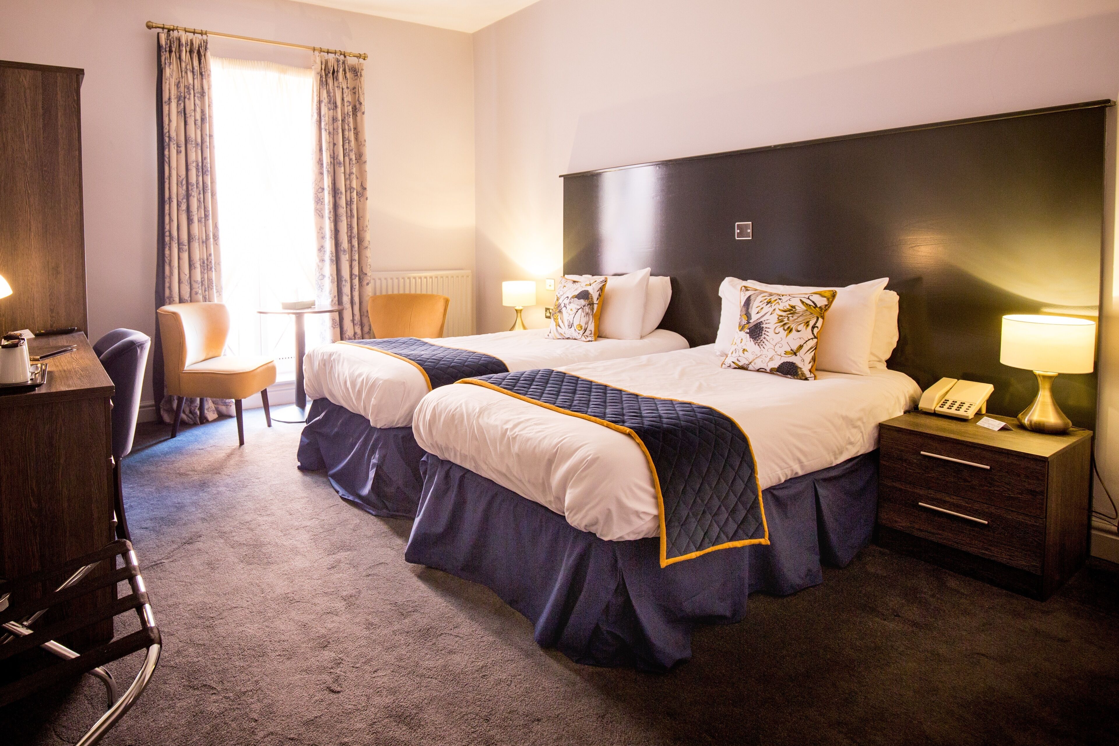 Een hotelkamer met twee bedden, een bureau, een stoel, een lamp en een raam met gordijnen.