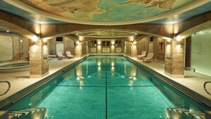 Une grande piscine intérieure avec un sol carrelé, entourée de chaises longues et agrémentée d’un plafond décoratif.
