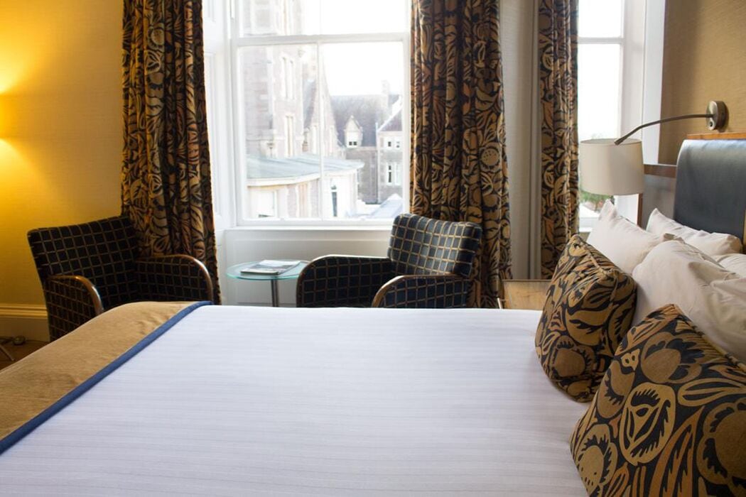A hotel room with a large bed, two armchairs, a small table, and a window with curtains.