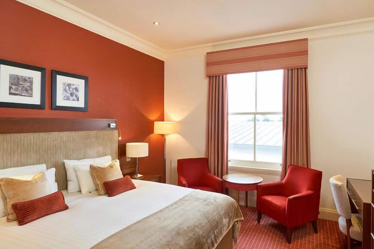A hotel room with a bed, two red chairs, a small table, a TV, and a window with curtains.