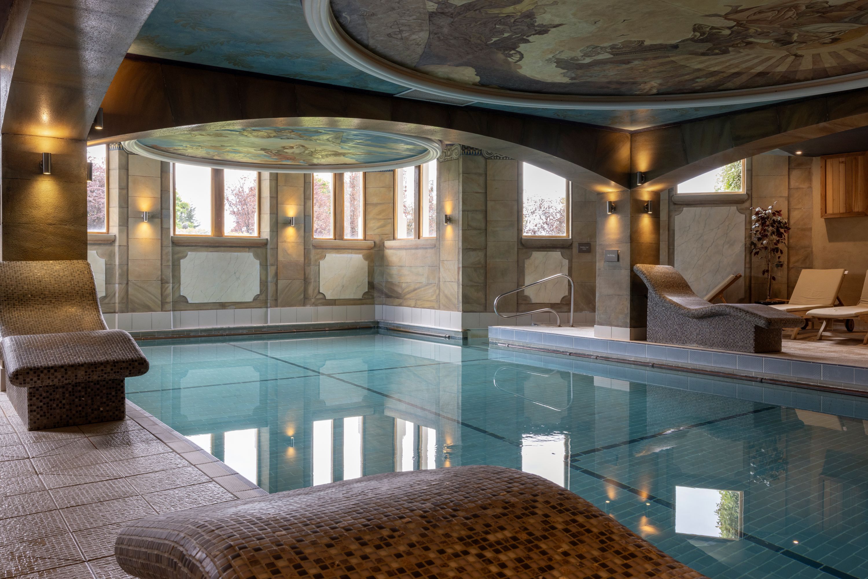 A large indoor swimming pool with clear water, surrounded by lounge chairs and a stone wall with windows.