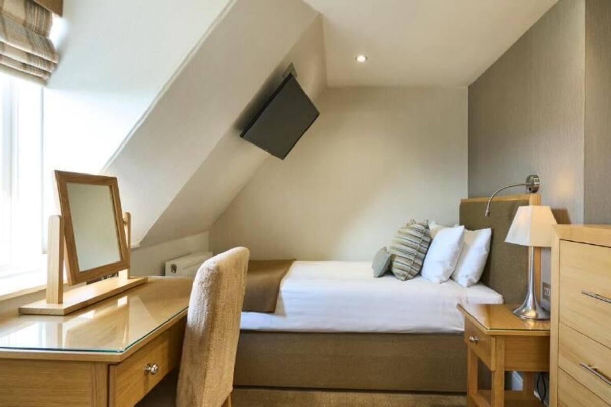 A modern bedroom with a sloped ceiling, a bed with pillows, a bedside table with a lamp, and a desk with a mirror.