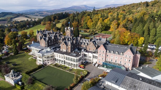Une vue aérienne d’un grand bâtiment historique à plusieurs ailes, entouré d’une végétation luxuriante et dominé par un paysage montagneux.