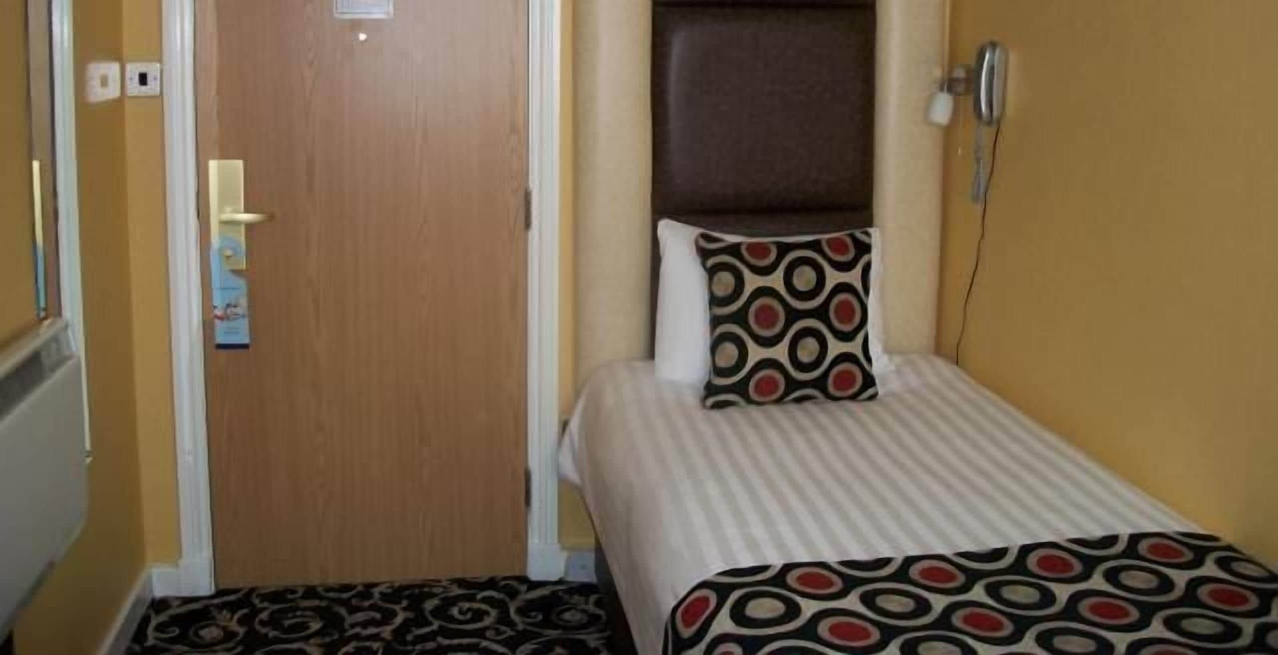 A small, single-bed room with a wooden door, a headboard, and a patterned bedspread.