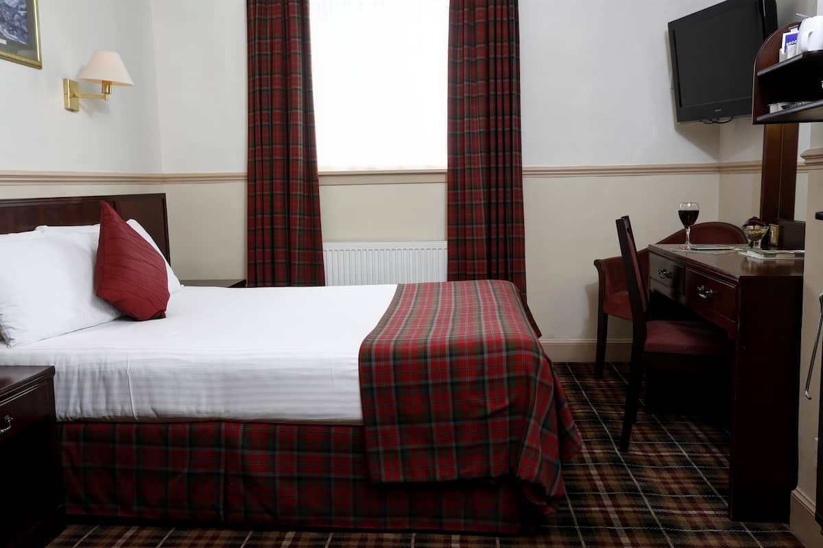 Quarto de hotel com cama, escrivaninha com cadeira, televisão e janela com cortinas.