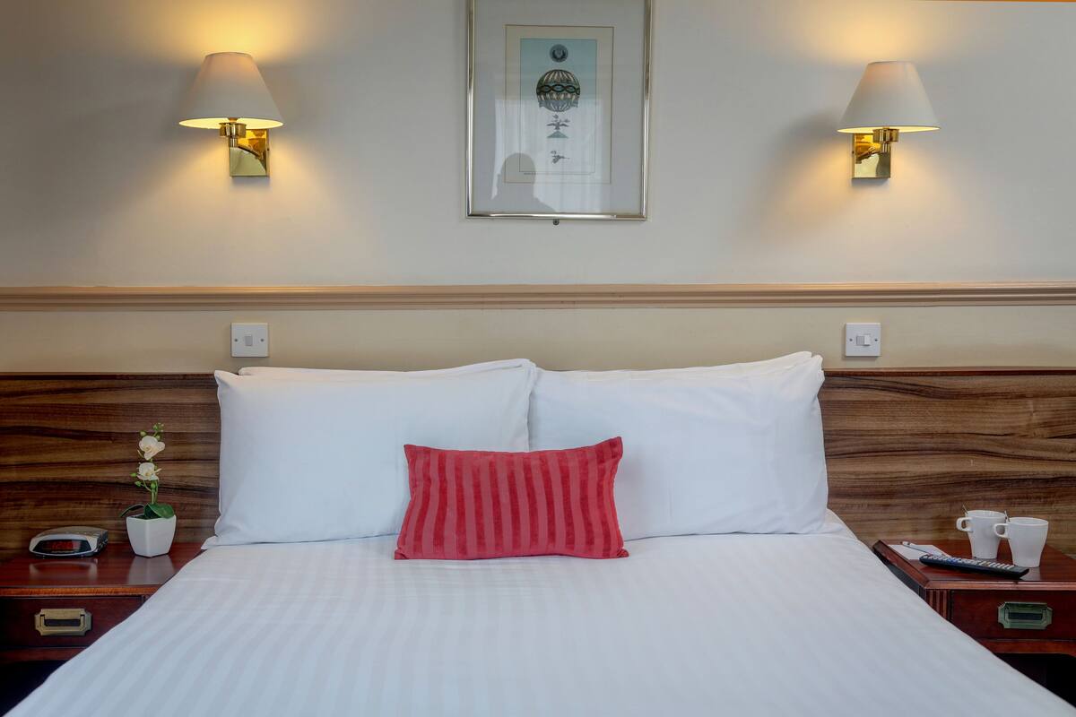 Cama bem arrumada com lençóis brancos, um travesseiro vermelho e duas mesas de cabeceira com luminárias e um quadro emoldurado acima.