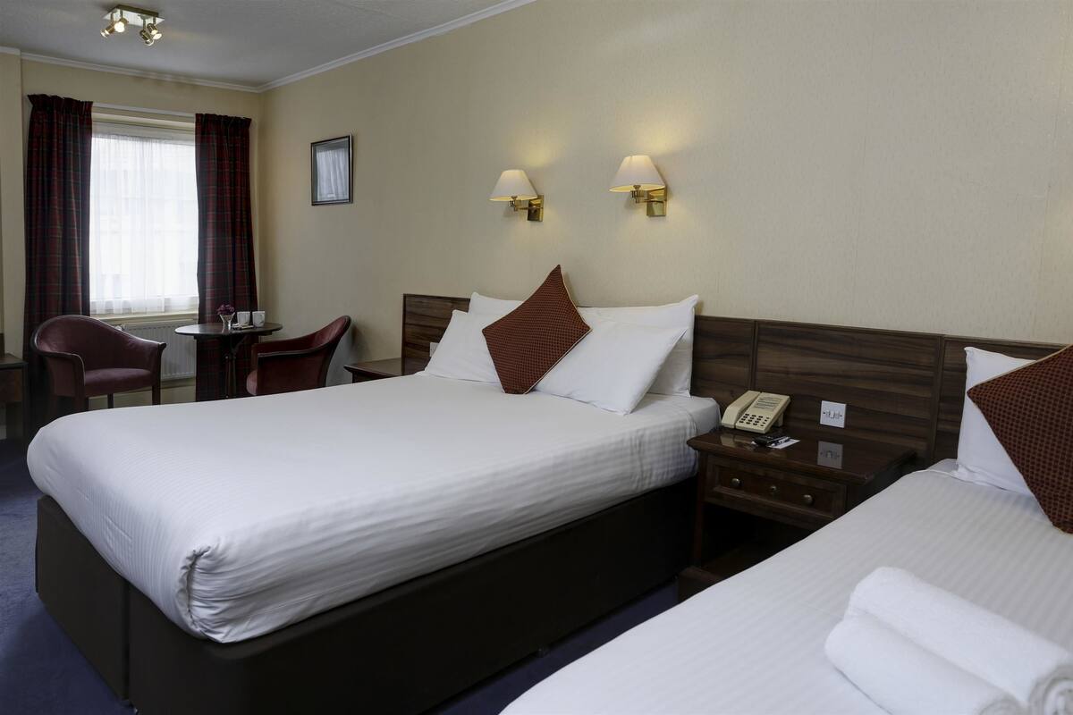 Quarto de hotel com uma cama grande, duas mesas de cabeceira, uma cadeira, uma mesa pequena e uma janela com cortinas.