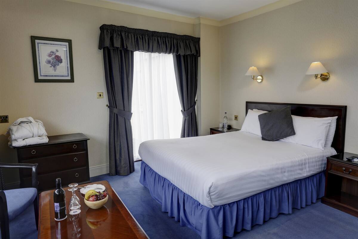 Quarto de hotel com uma cama grande, mesa de cabeceira de madeira, escrivaninha com uma garrafa e copos, uma fruteira e um quadro na parede.