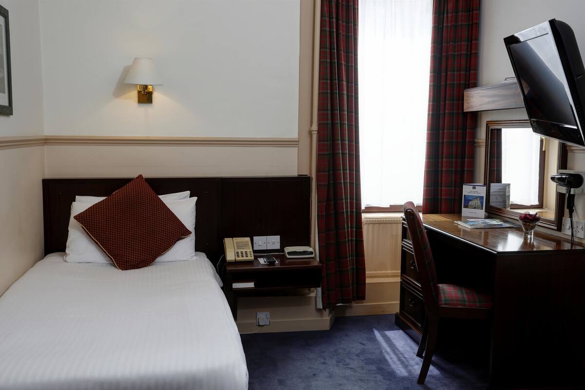 Quarto de hotel com uma cama, uma escrivaninha com computador, telefone e televisão.