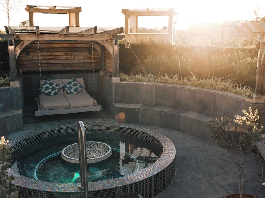A hot tub with a wooden deck and seating area, surrounded by stone walls and plants.