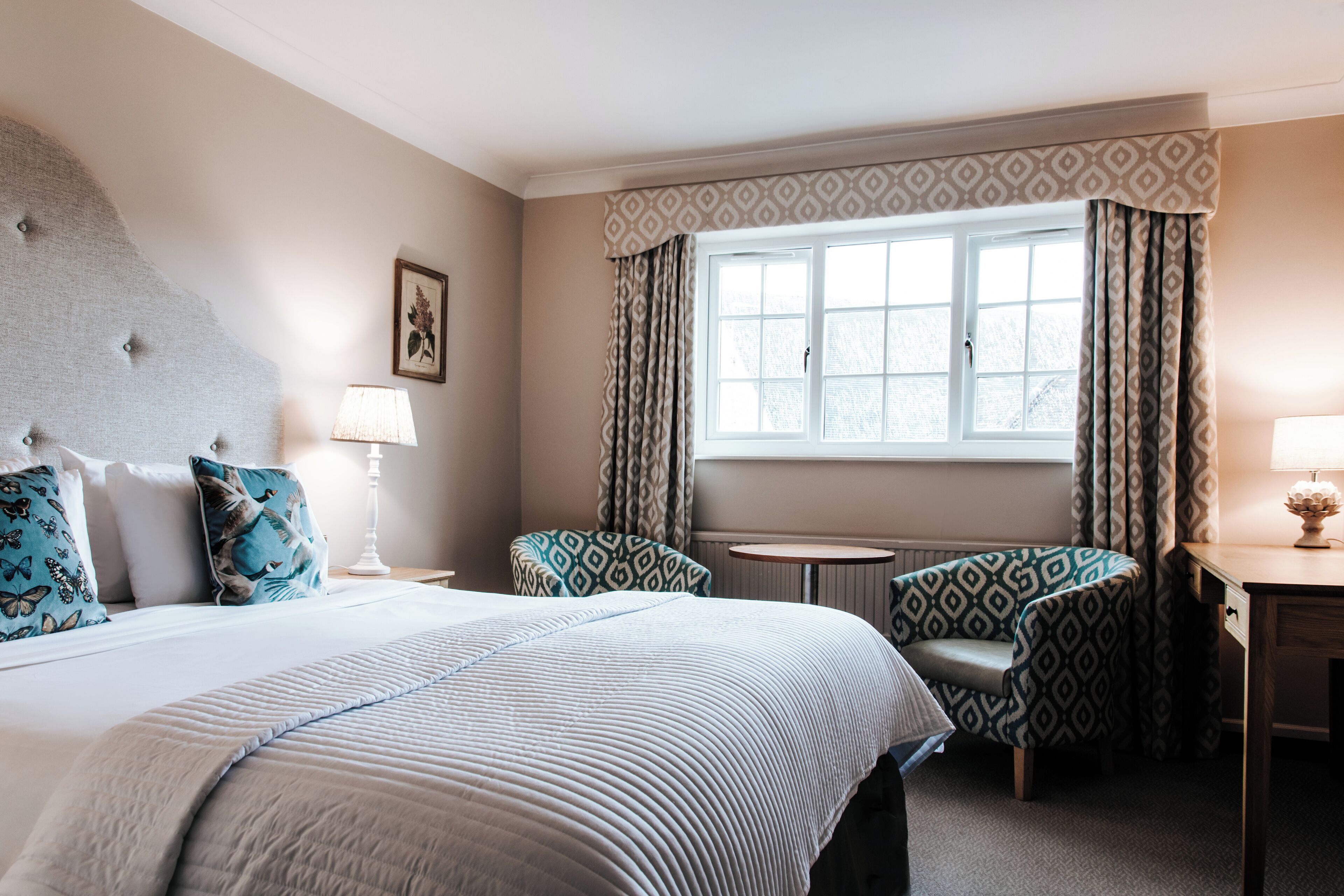 Une chambre d’hôtel avec un grand lit, deux fauteuils à motifs, une petite table ronde et une fenêtre avec des rideaux.