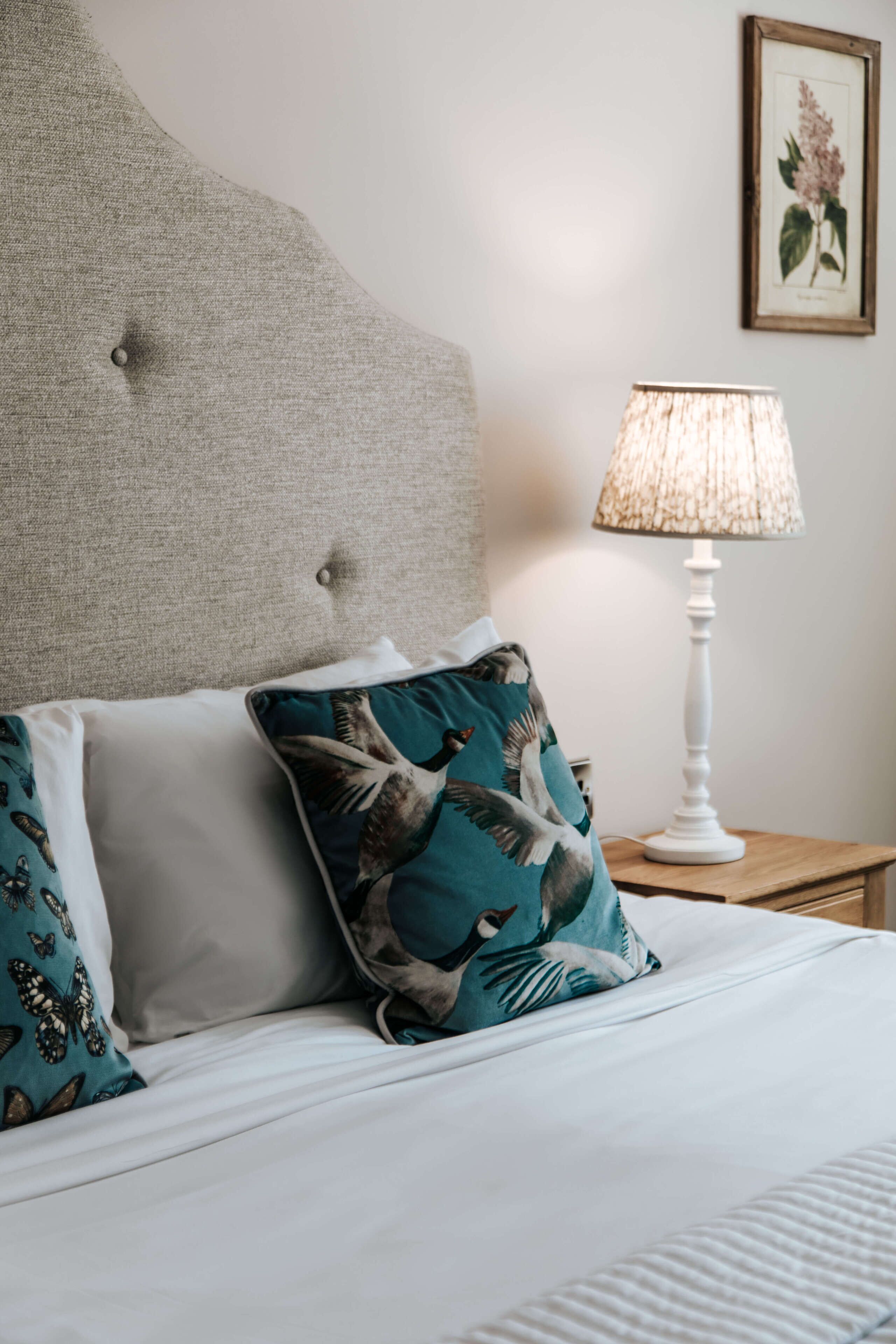 A neatly made bed with a tufted headboard, two pillows with a bird pattern, and a white lamp on a wooden nightstand.
