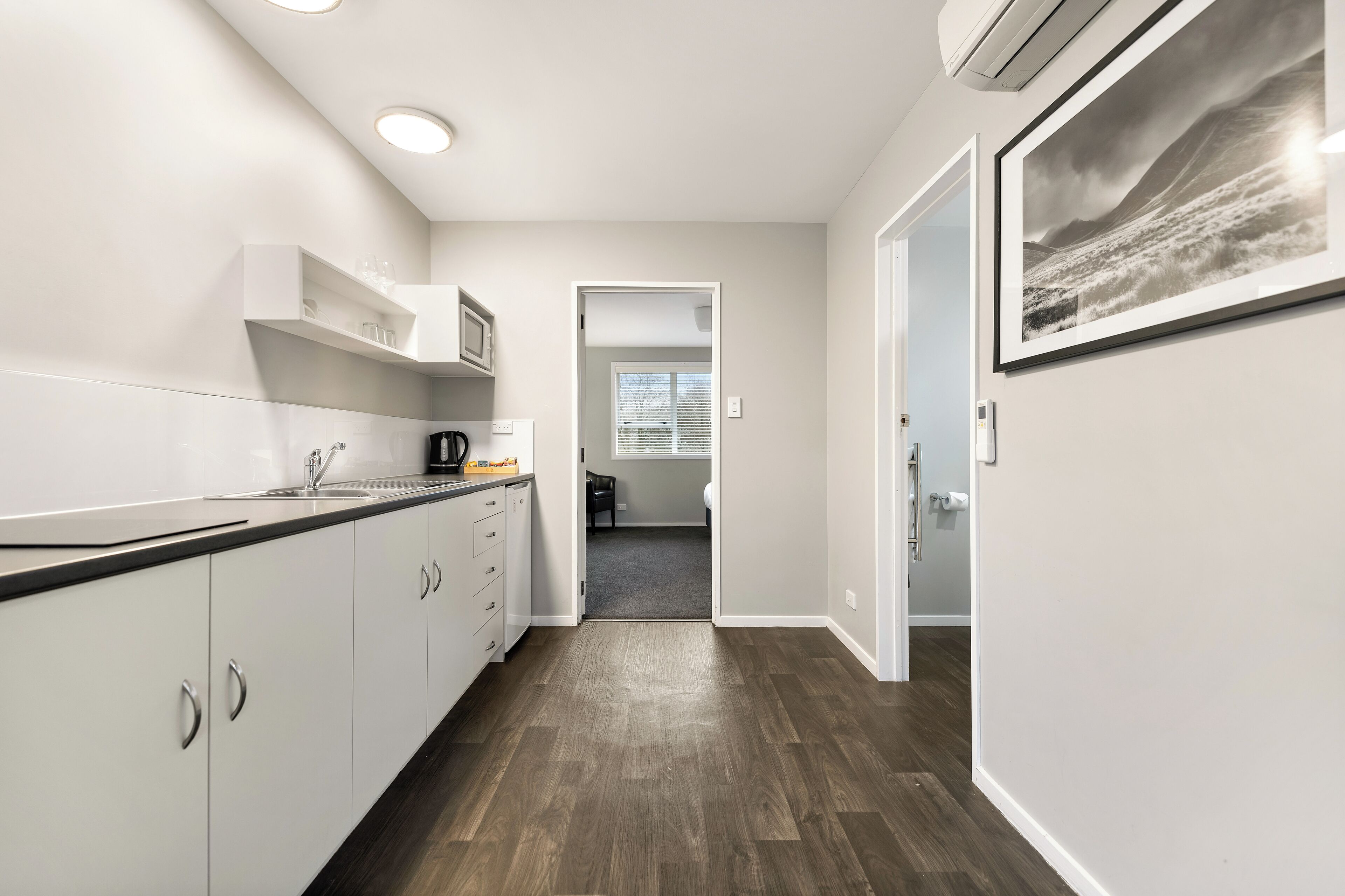 A modern kitchen with white cabinets, a black countertop, and a view into a bedroom through an open door.