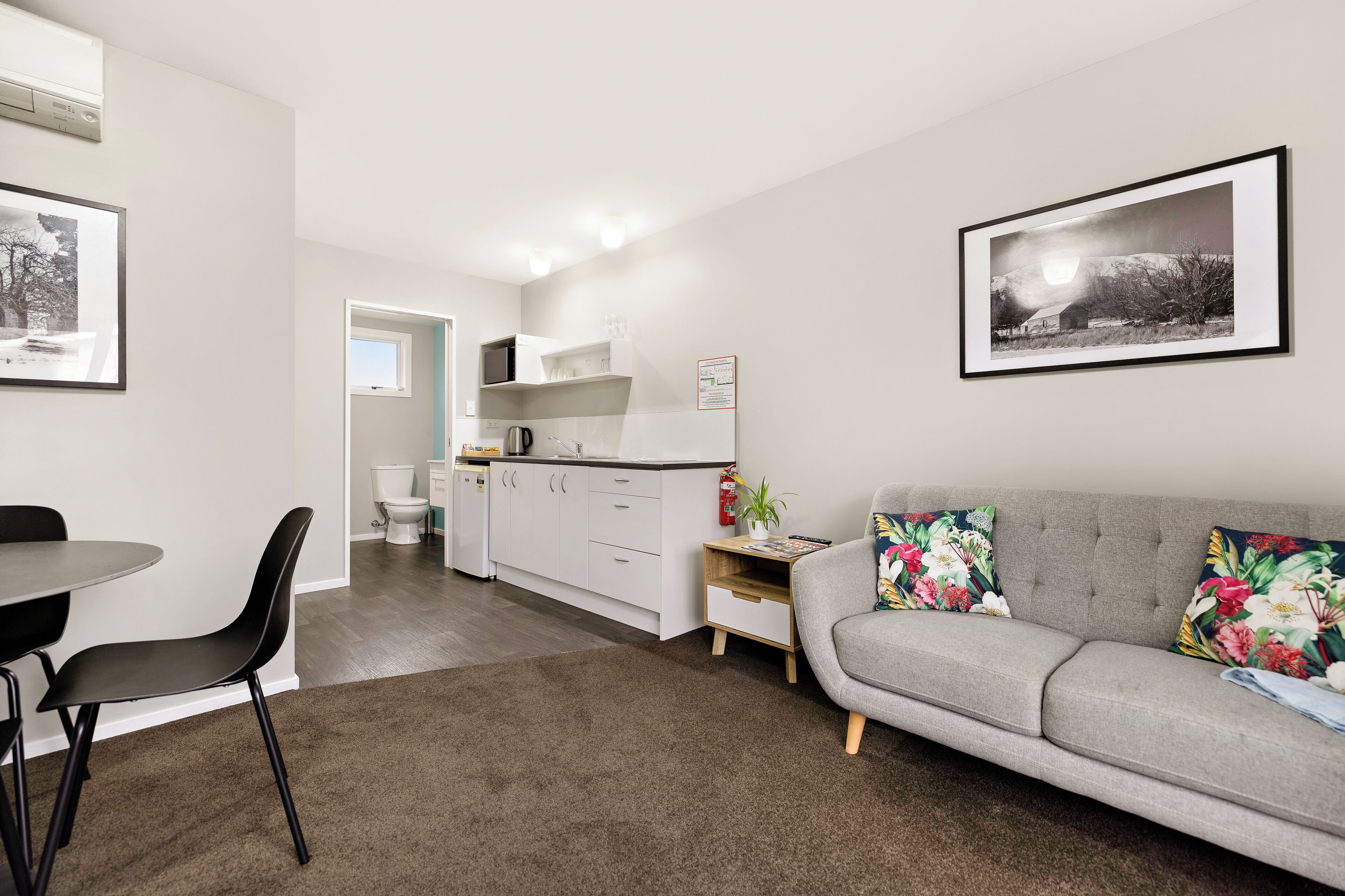 A modern living room with a grey sofa, a dining table with chairs, a kitchen area with white cabinets, and a framed photograph on the wall.