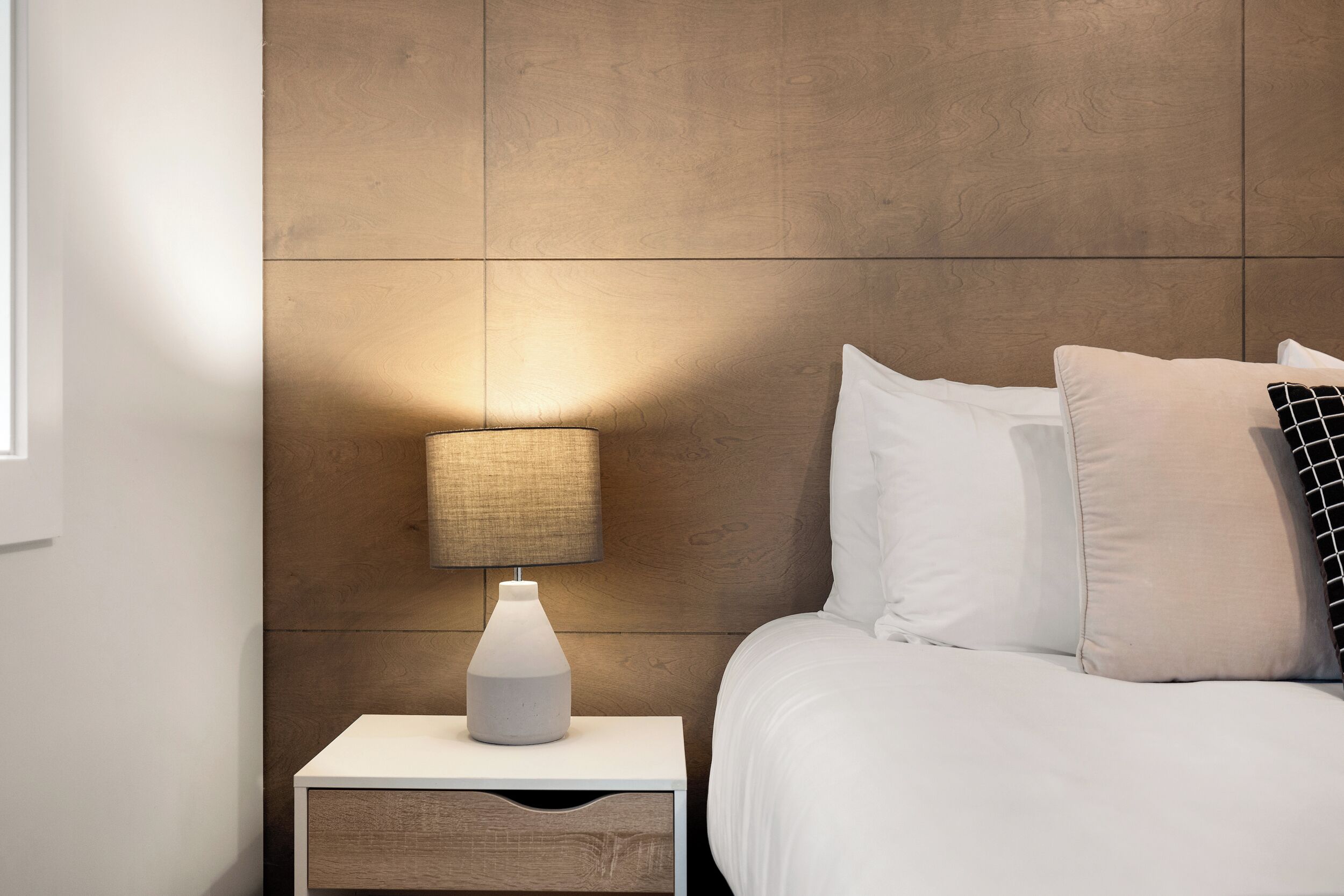 Uma cama bem arrumada com roupa de cama branca, um abajur e uma mesinha de cabeceira com uma gaveta.