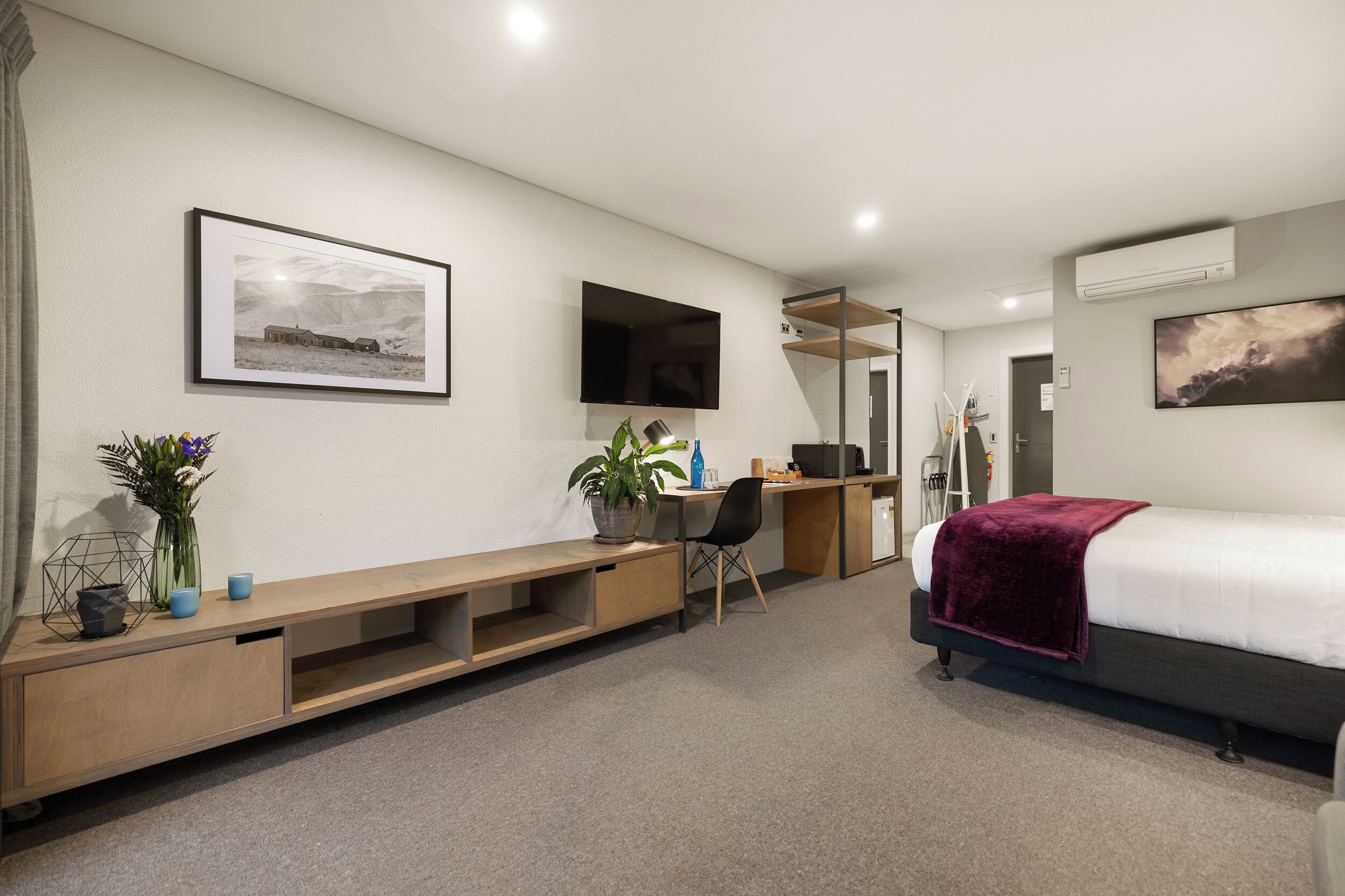 A modern hotel room with a bed, desk, and seating area. There are two framed pictures on the walls, a potted plant, and a small table with a lamp.