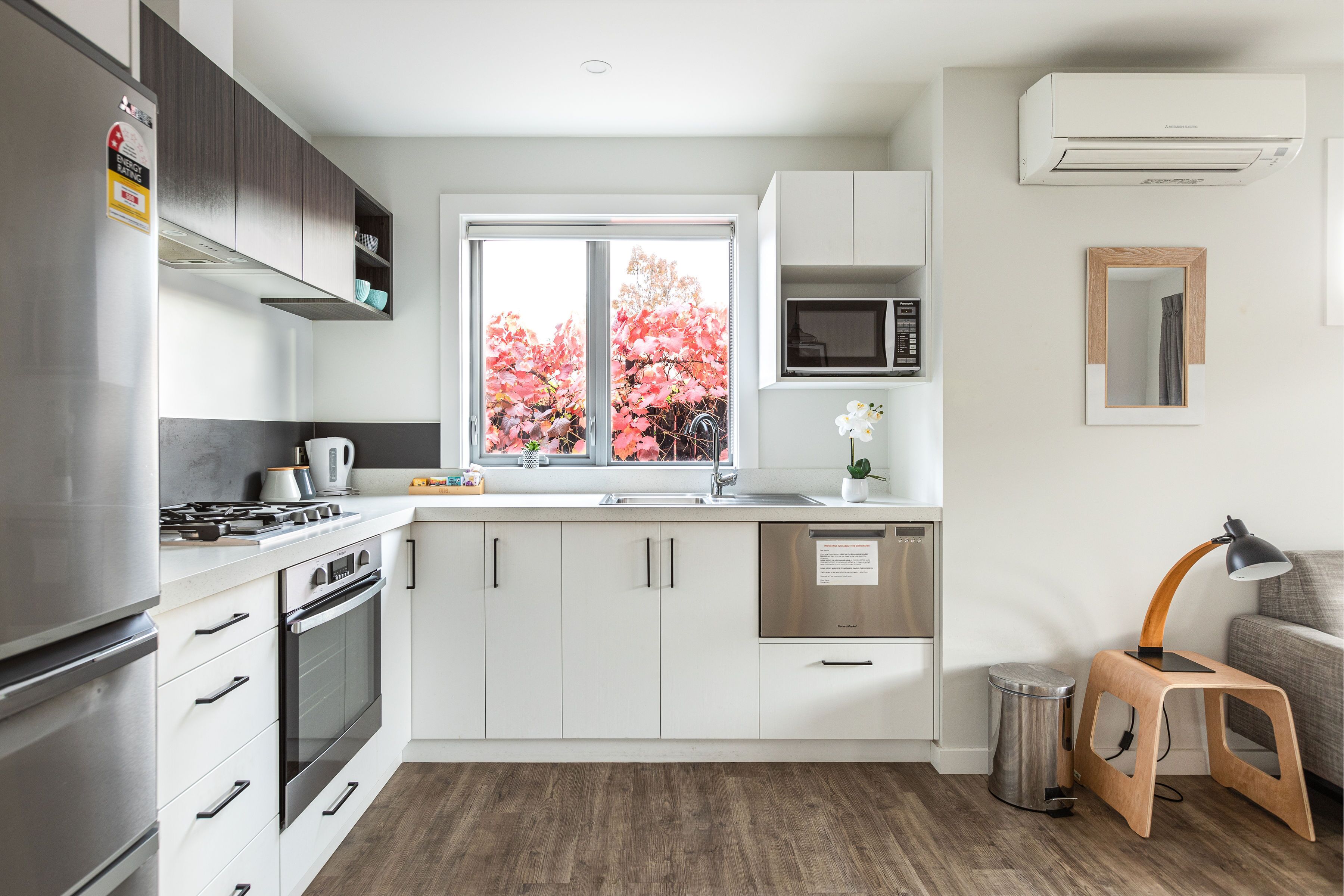 A modern kitchen with white cabinets, stainless steel appliances, a window with a view of pink trees, and a small stool with a lamp.