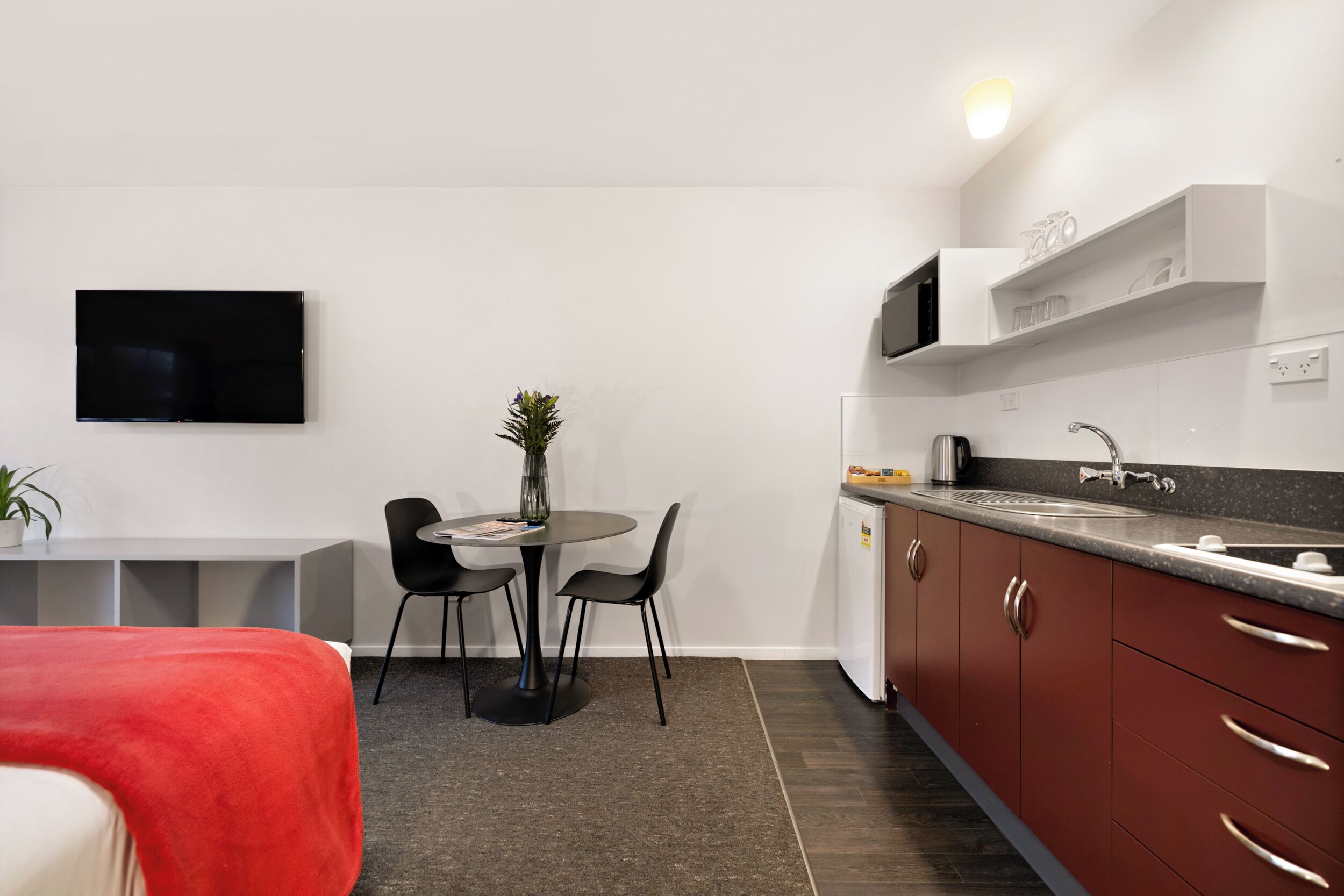 A compact hotel room with a kitchenette, a dining area with a small table and chairs, a flat-screen TV mounted on the wall, and a red bedspread.