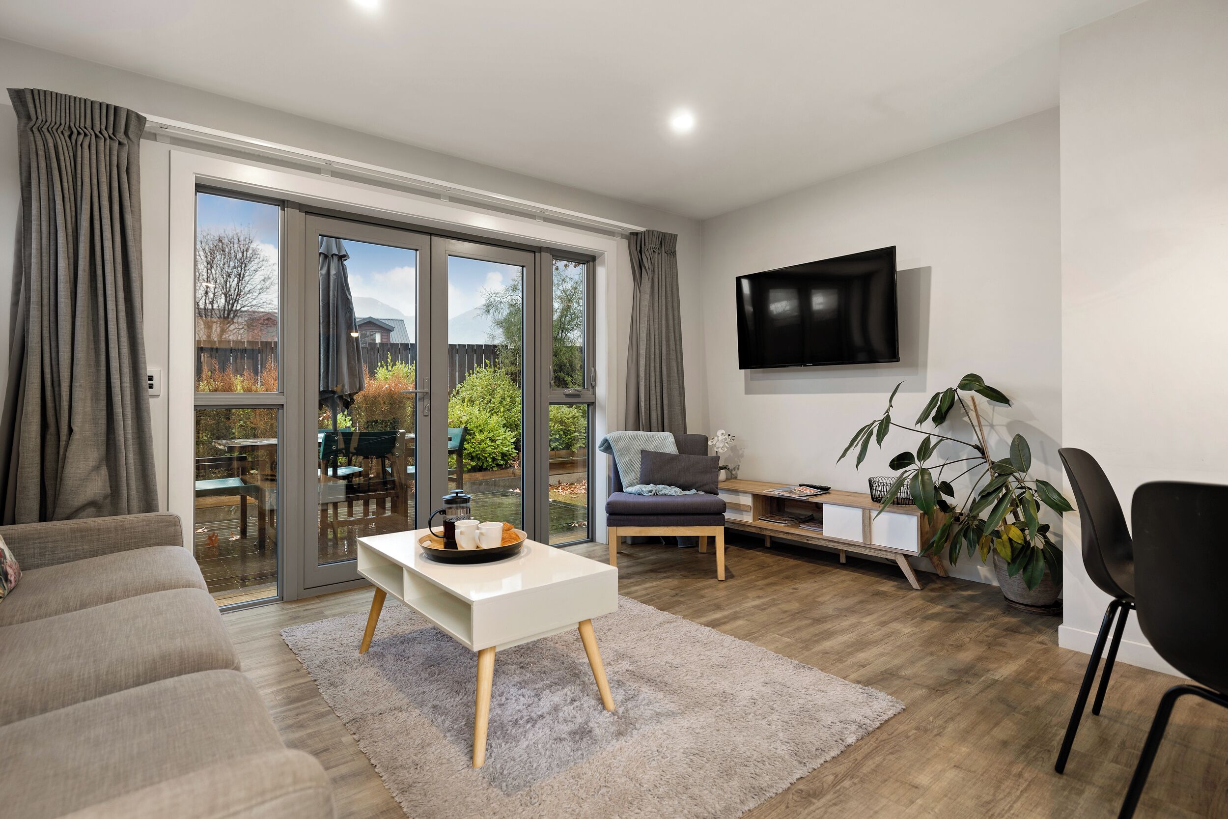 A modern living room with a sofa, a coffee table, a TV mounted on the wall, and a sliding glass door leading to an outdoor area.