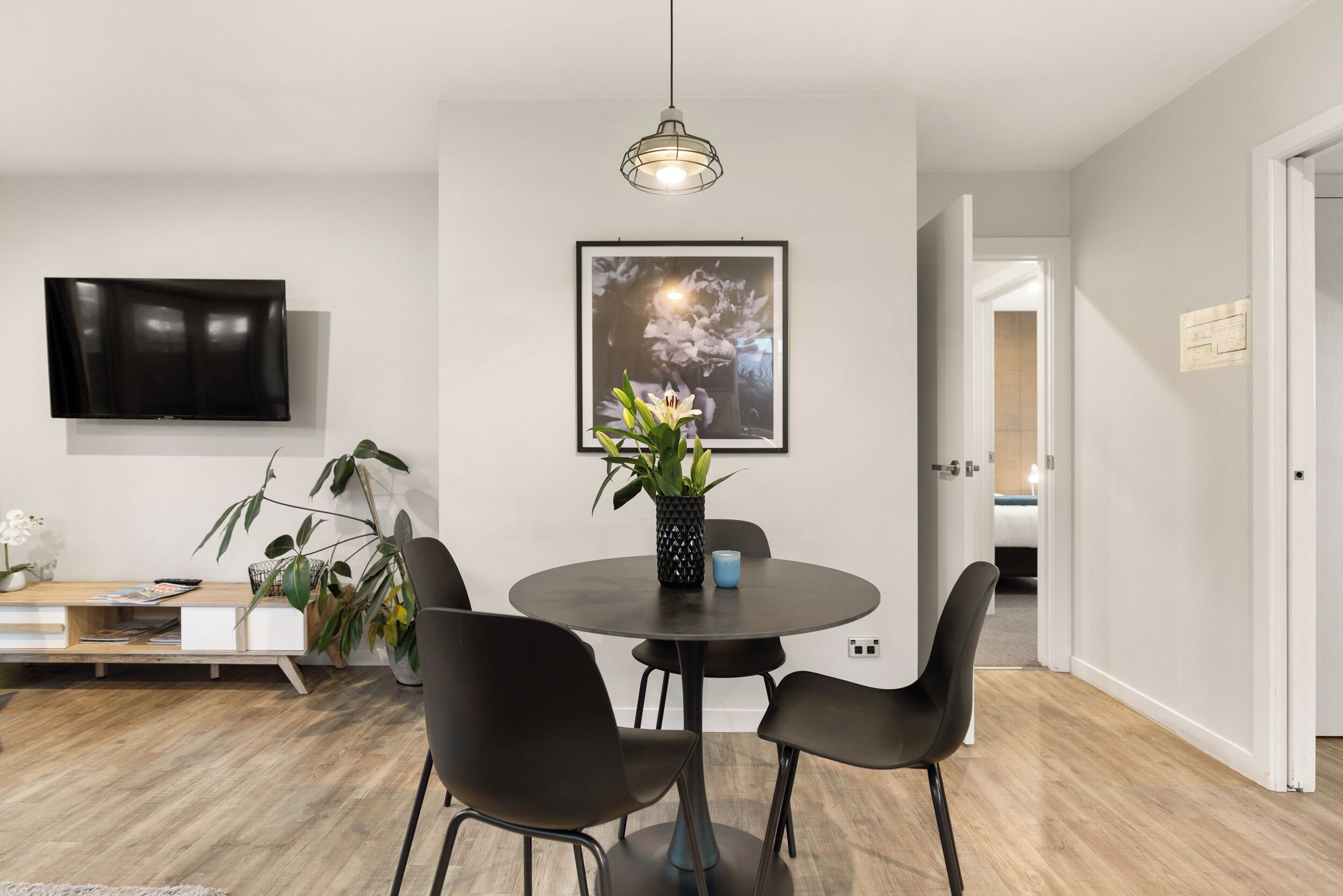 A modern dining area with a round table, black chairs, a vase with flowers, a framed picture on the wall, and a television mounted above.