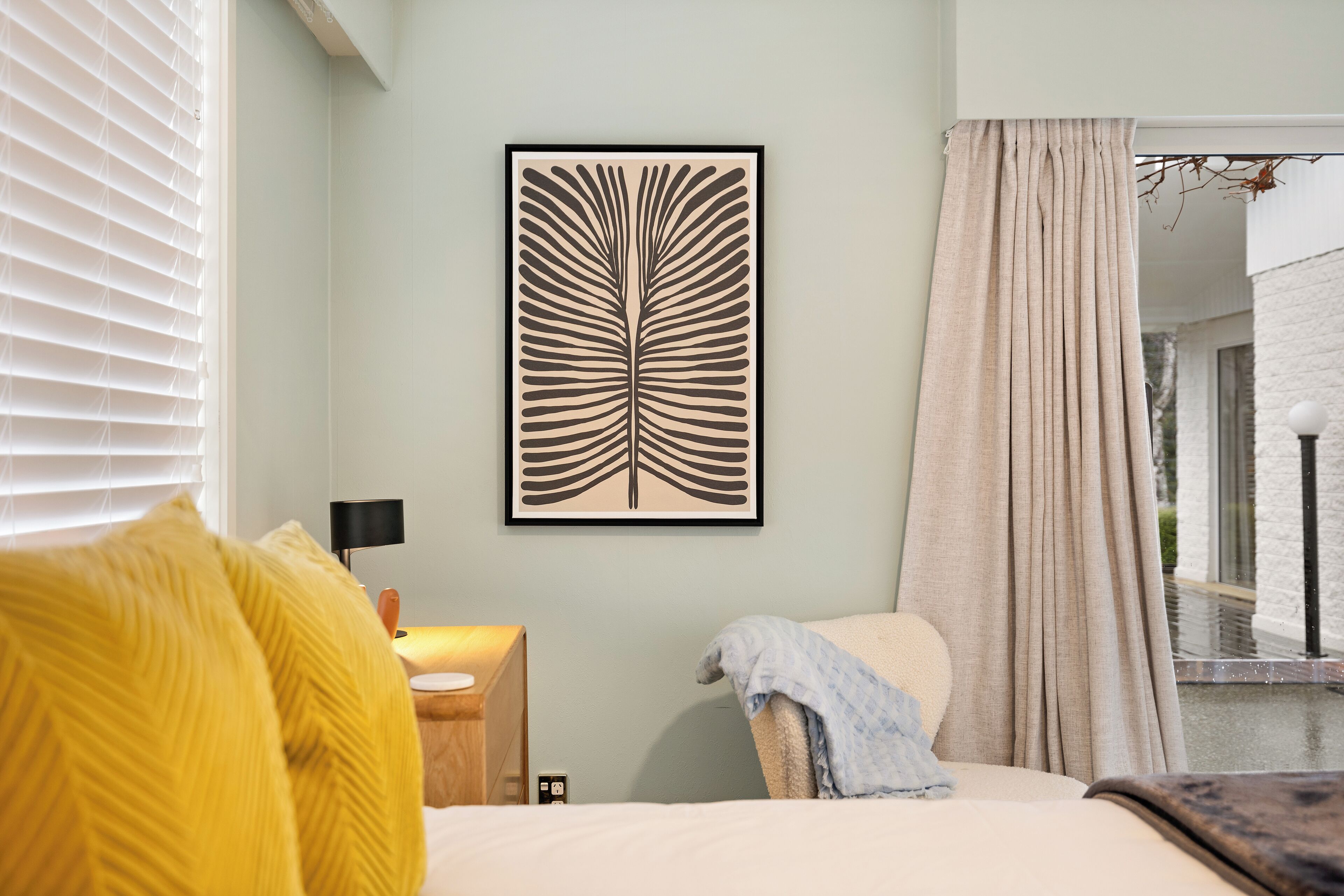 Un dormitorio con una cama, una mesita de noche con lámpara, una ventana con persianas, una cortina y una obra de arte enmarcada en la pared.