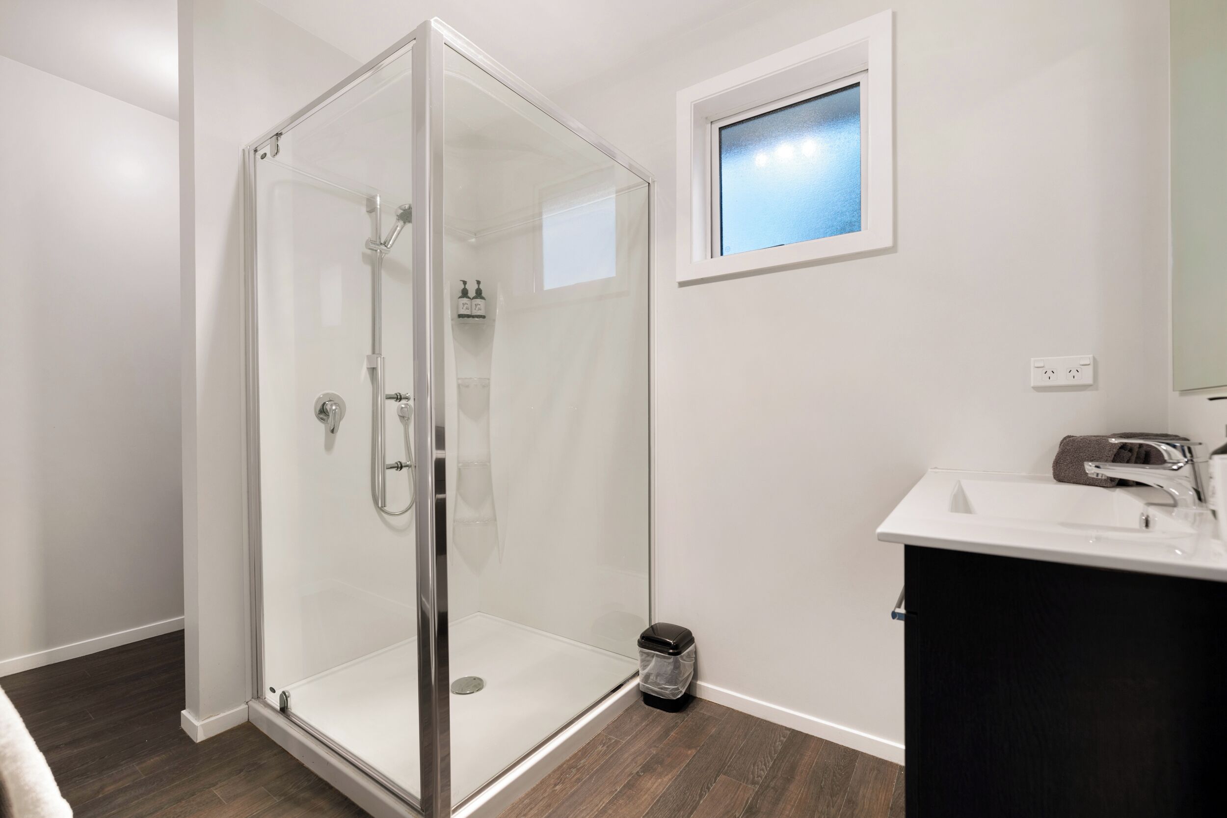 Ein modernes Badezimmer mit einer Duschkabine aus Glas, einem weißen Waschbecken und einem kleinen Fenster darüber.