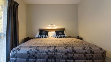 A neatly made bed with a floral-patterned bedspread, two pillows with text, a bedside lamp, and a window with curtains.
