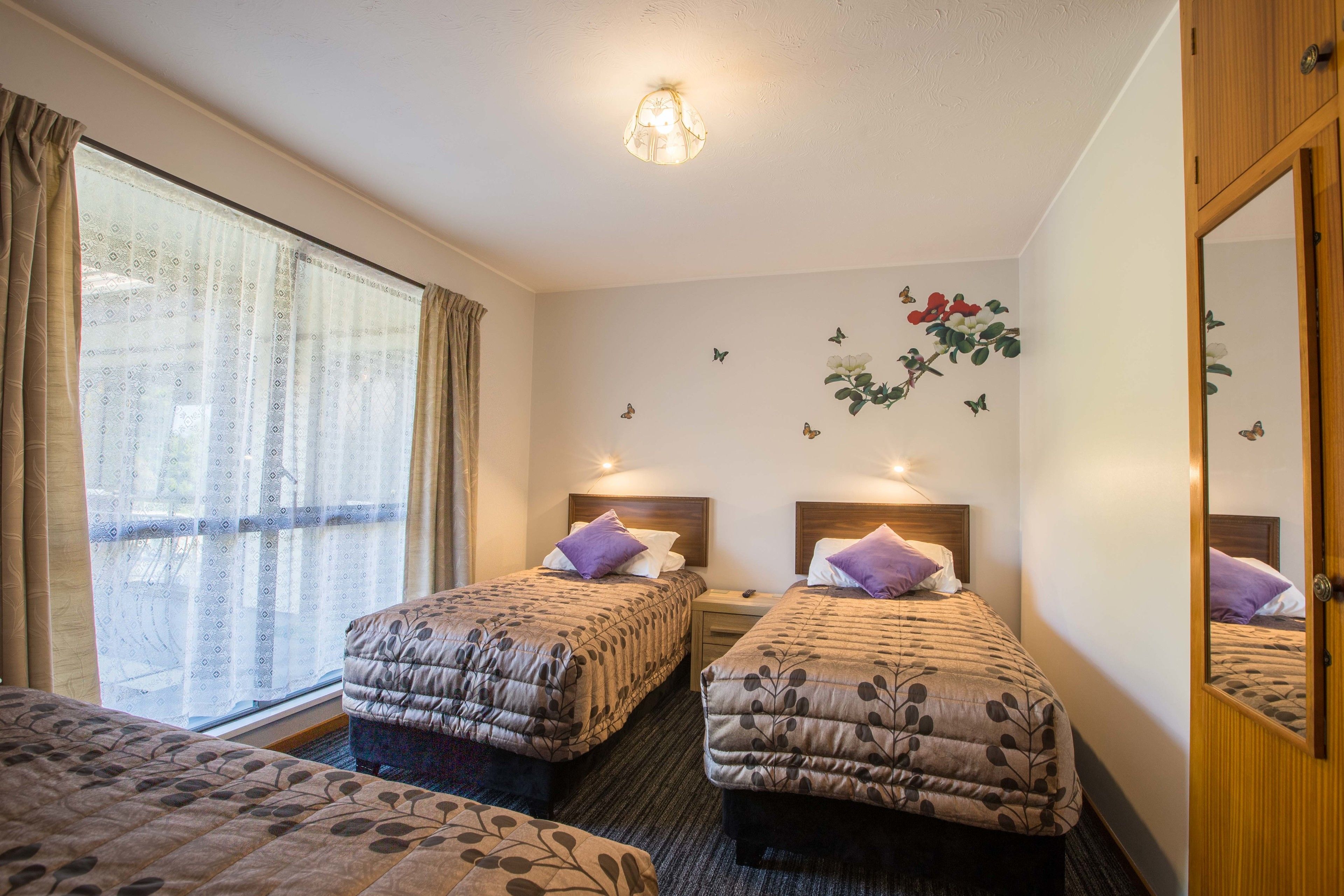 A hotel room with two beds, a window with curtains, a ceiling light, and wall decorations including butterflies and flowers.