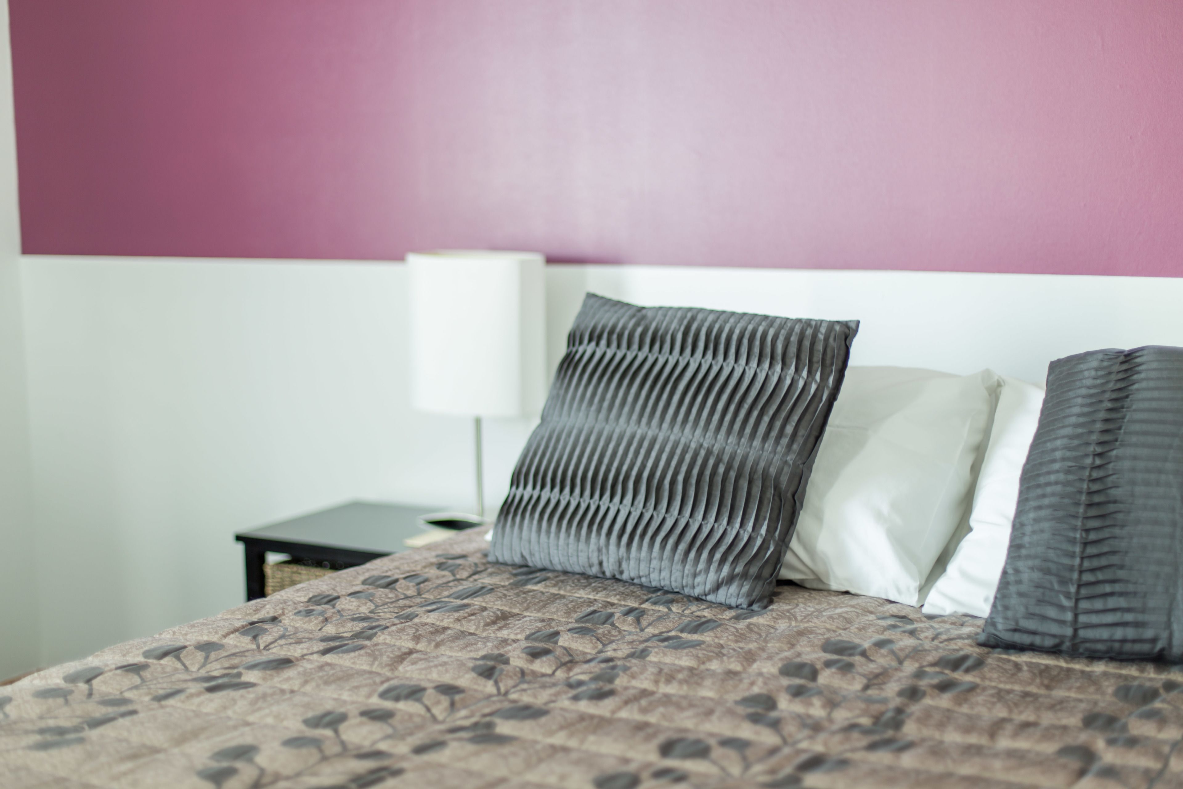 Uma cama com colcha estampada, duas almofadas, uma mesinha de cabeceira e um abajur em frente a uma parede rosa e branca.