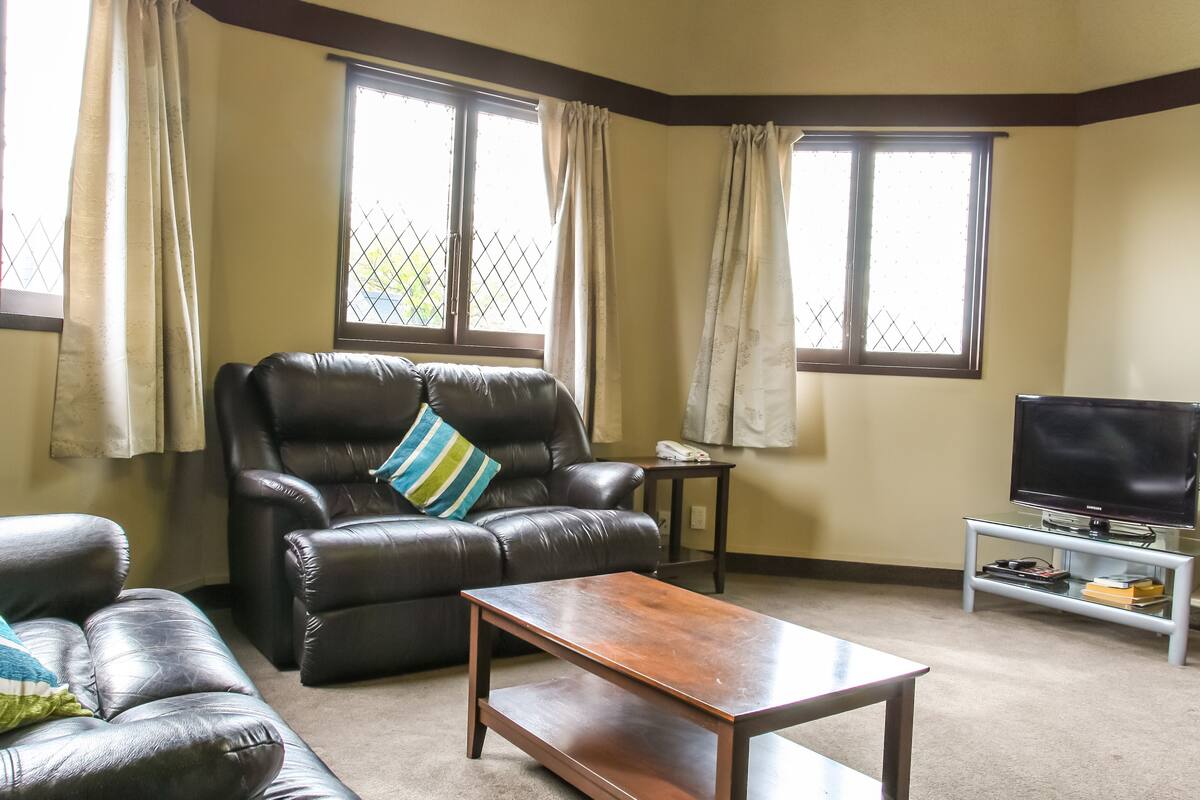 Ein Wohnzimmer mit einer Ledersofa, einem Holztisch, einem Fernseher auf einem Standfuß und großen Fenstern mit Vorhängen.