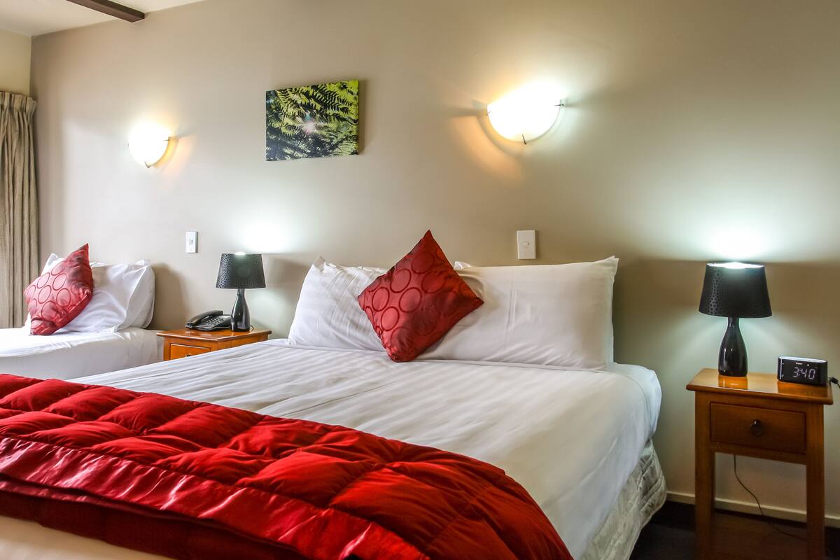 Ein Hotelzimmer mit zwei Betten, roter und weißer Bettwäsche, einer roten Tagesdecke, zwei Nachttischen mit Lampen, einer Uhr und einer wandmontierten Leuchte.