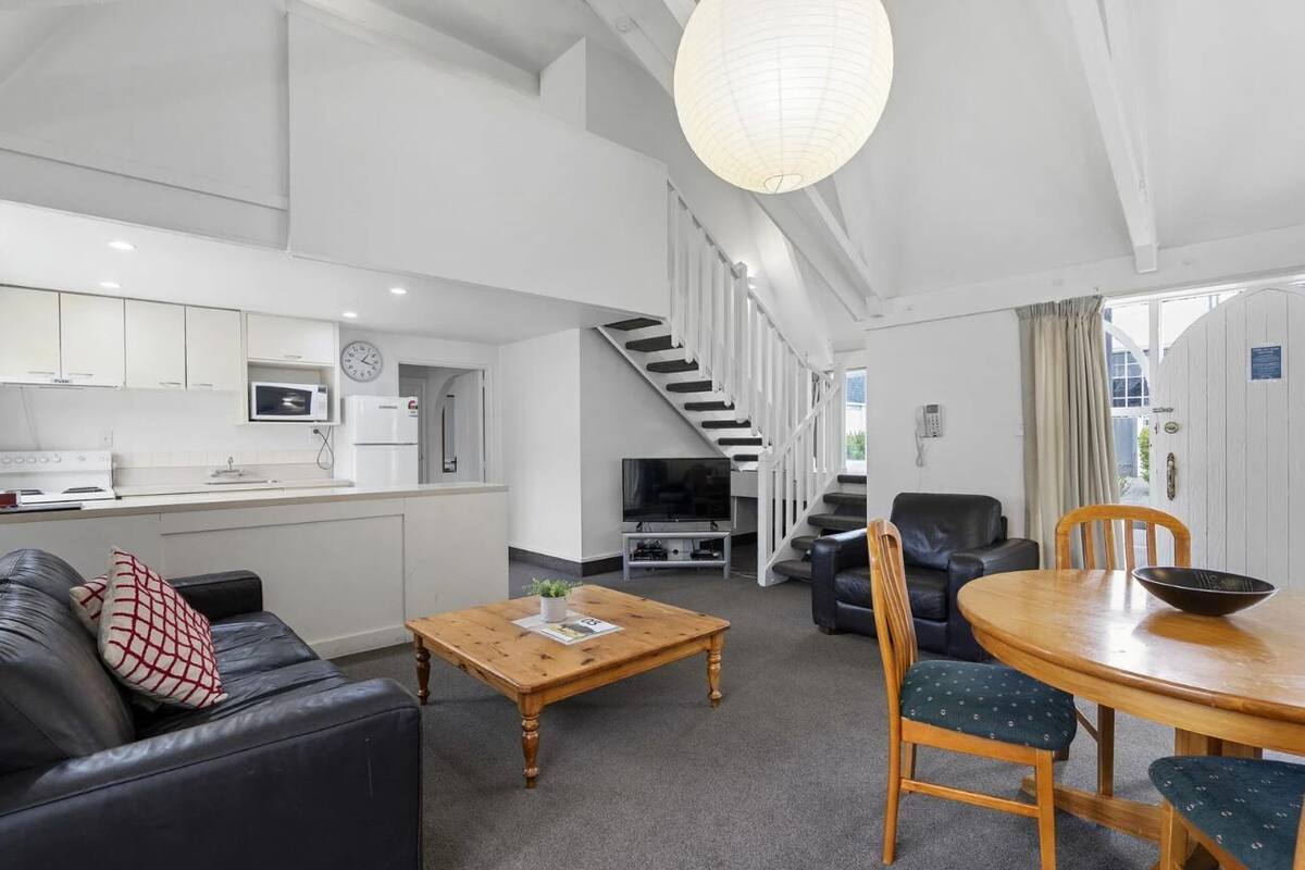 Ein modernes Wohnzimmer mit Sofa, Couchtisch und Esstisch. Es gibt eine Küchenzeile mit Schränken und Geräten, sowie eine Treppe zu einer weiteren Ebene.