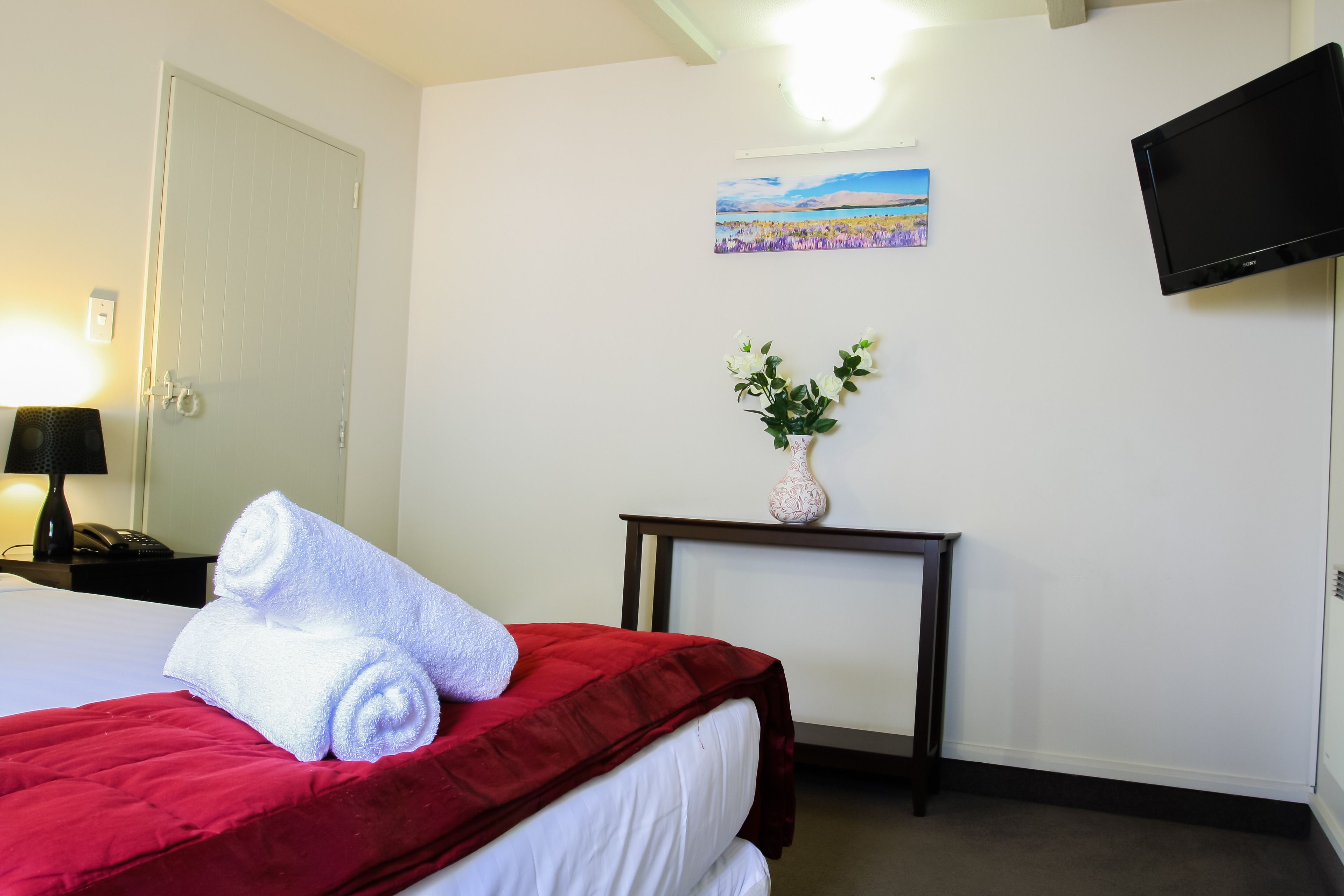 Una habitación de hotel con una cama, un edredón rojo, dos toallas enrolladas, una mesita con un jarrón, un televisor de pared y un cuadro sobre el televisor.