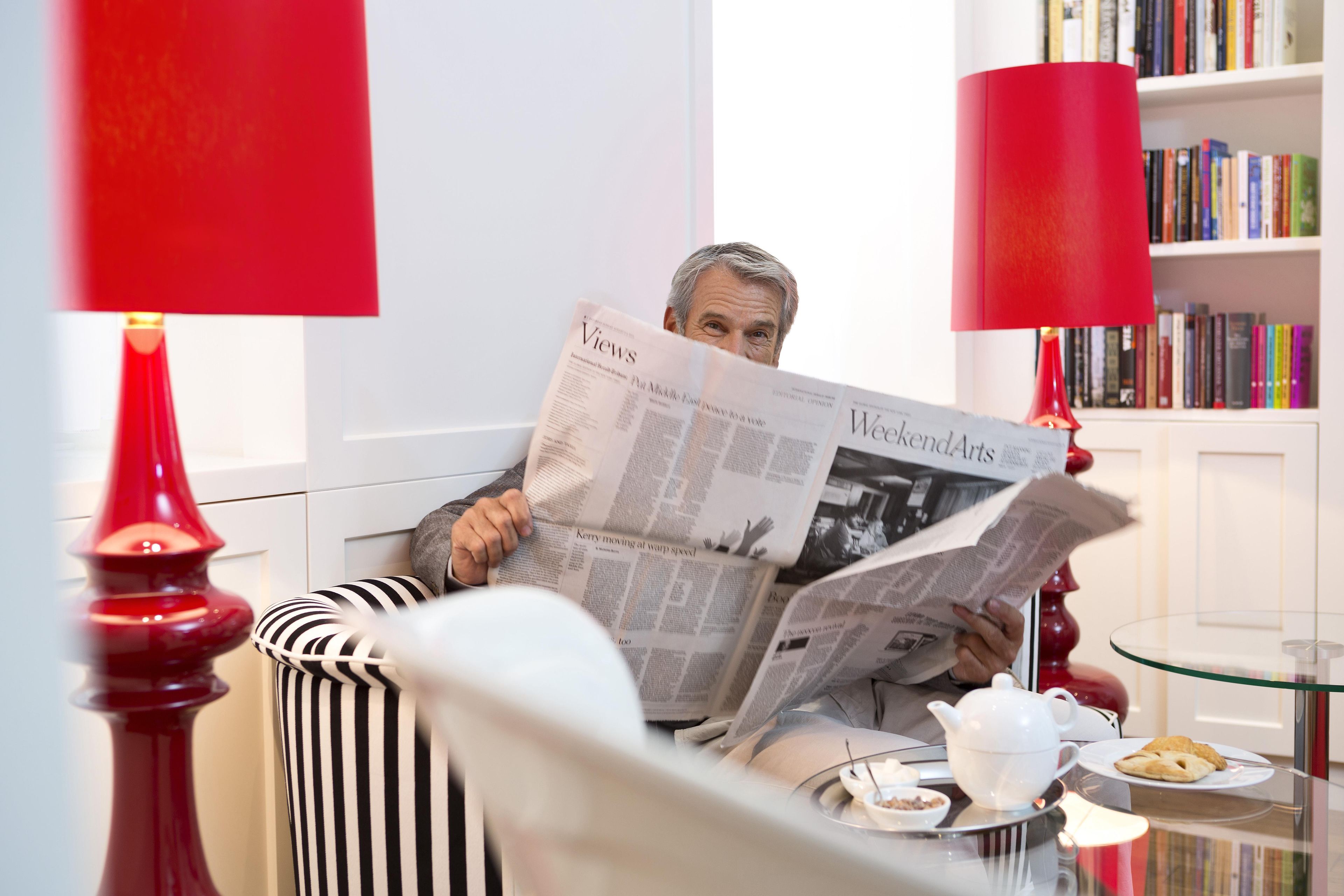 Una persona leyendo un periódico en una habitación con una silla a rayas, una mesa de vidrio y una estantería al fondo.