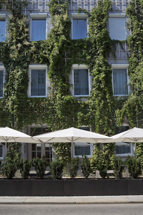 A building with multiple windows covered in ivy, outdoor seating with white umbrellas, and small potted plants in front. - The Harmonie Vienna, BW Premier Collection (Vienna)