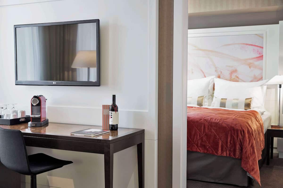 A hotel room with a bed, a desk with a coffee maker, a bottle of wine, a chair, and a TV on the wall.