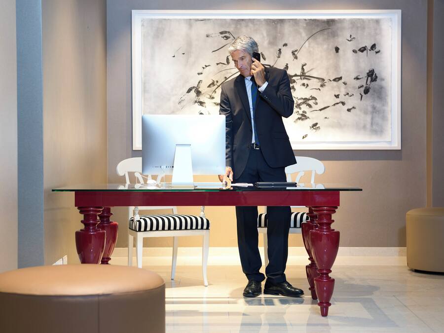 A man in a suit standing at a glass table with a laptop, a phone, and papers. There is a chair and ottoman in the foreground, and a wall with a bird illustration in the background.