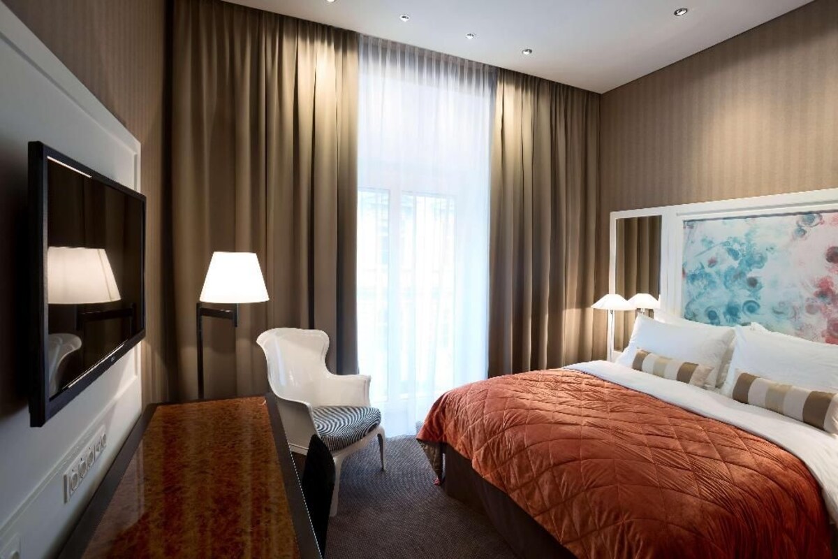 Habitación de hotel con cama, una silla, escritorio, televisor y un ventanal con cortinas.