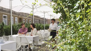 Un café al aire libre con mesas y sillas, un camarero pasando y un edificio al fondo.