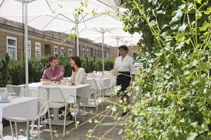 Un café al aire libre con mesas y sillas, un camarero pasando y un edificio al fondo.