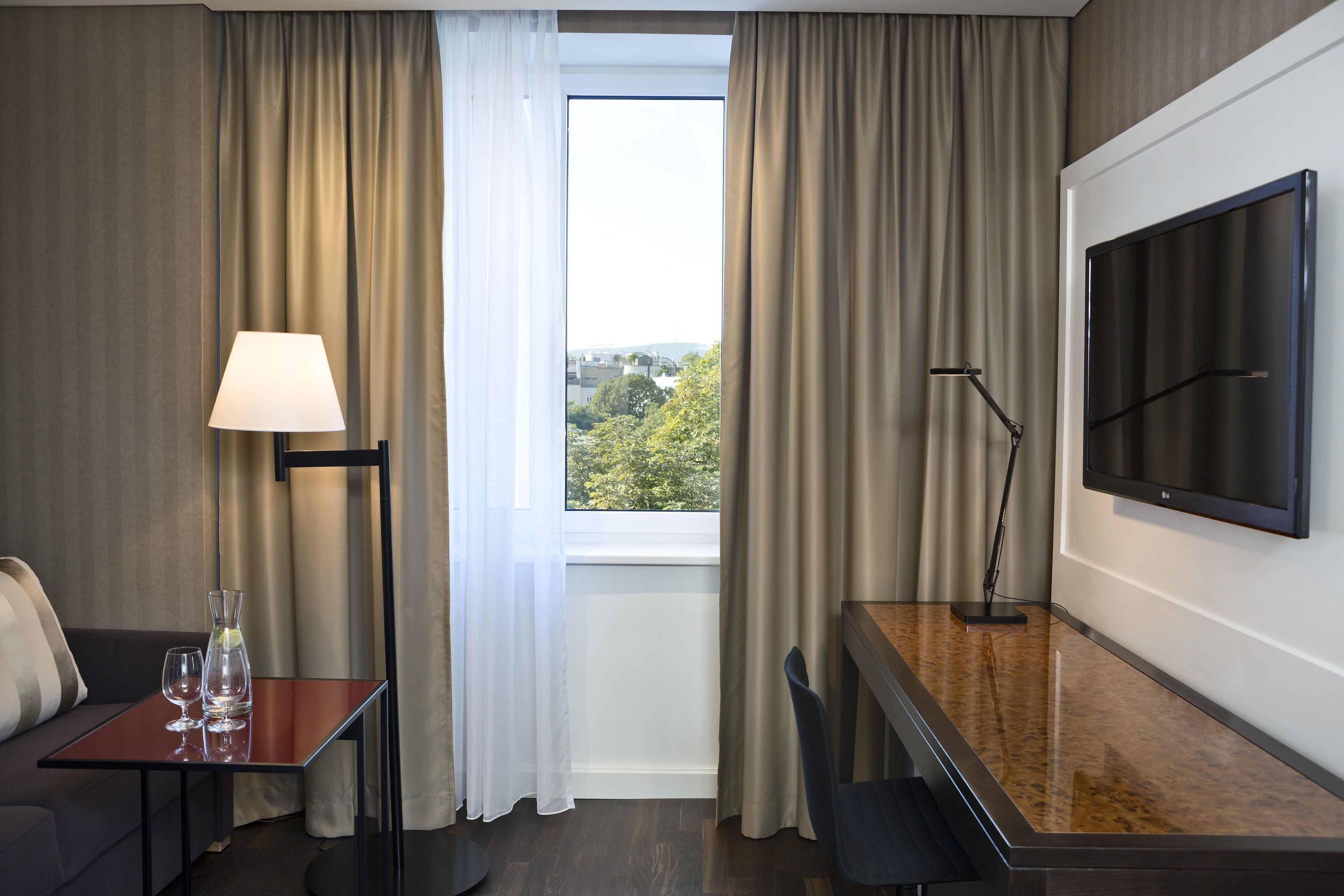 A hotel room with a bed, a desk with a chair, a TV, a lamp, and a window with curtains.